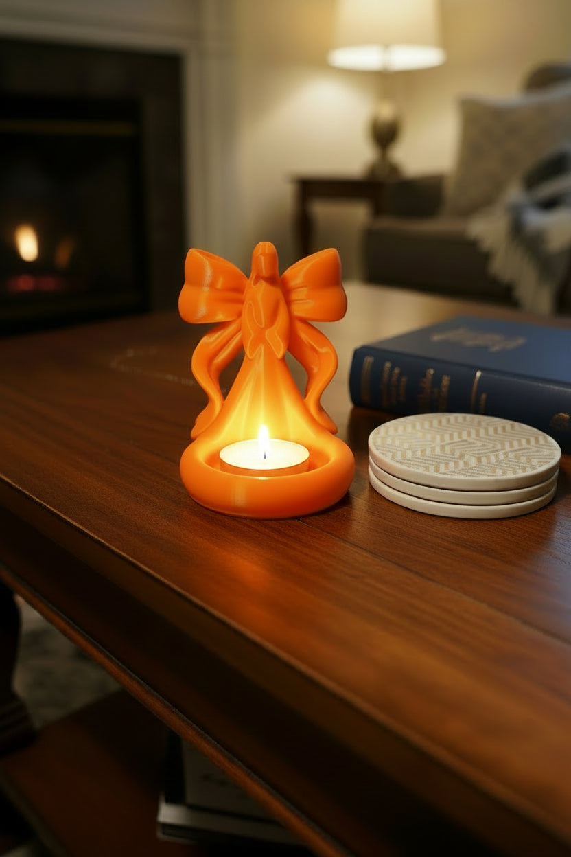 Sunset Orange 3D-printed tea light holder with an angelic figure detailed with bow style ribbon wings glowing on a table next to coasters and a book.