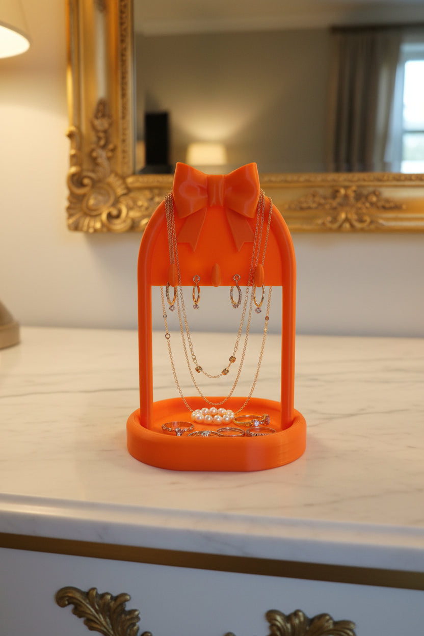 Sunset Orange 3D-printed jewelry stand holding necklaces and earrings with rings in bottom tray crowned with a bow on a vanity.