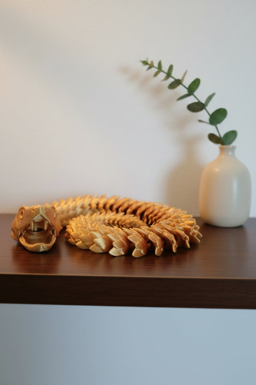 Gold Silk 3D-printed articulated viper coiled with open jaw showing fangs and dragon type scales on a wood shelf by a plant.