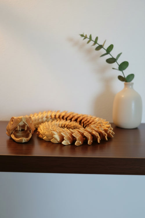 Gold Silk 3D-printed articulated viper coiled with open jaw showing fangs and dragon type scales on a wood shelf by a plant.