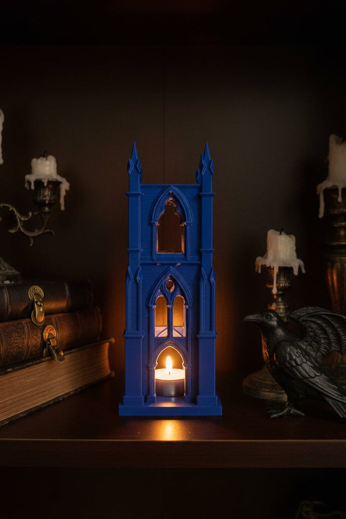 Cobalt Blue 3D-printed tea light holder with towering cathedral design and gothic arched windows glowing on a shelf next to a crow and gothic decor.